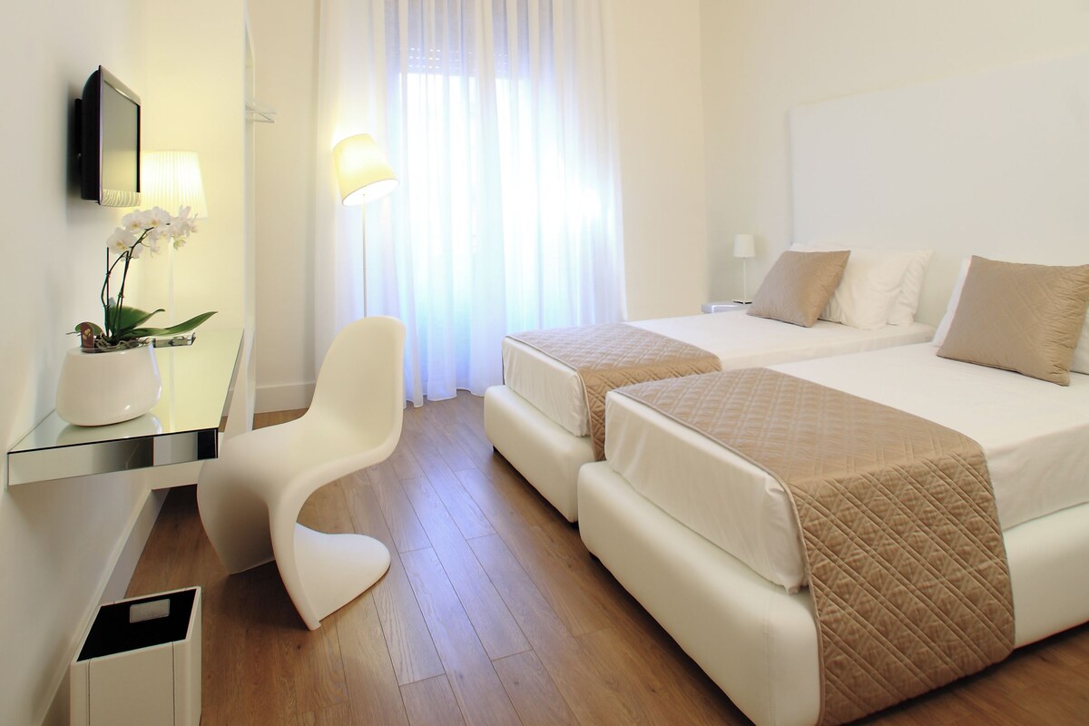 A hotel room with a wooden floor, two beds, a desk with a chair, a mirror, and a ceiling with decorative patterns.