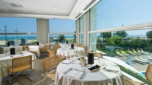 A restaurant with round tables set for dining, large windows offering a view of the beach and ocean, and outdoor seating with lounge chairs. - Hotel Sarti (Riccione)