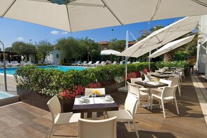 Un área junto a la piscina con sillones de descanso, una piscina y mesas de comedor al aire libre con sombrillas.