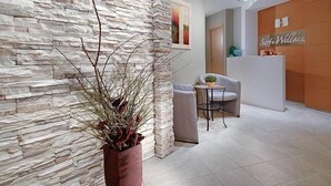 A reception area with a stone wall, a reception desk, a small round table with chairs, and a potted plant. - Hotel Sarti (Riccione)