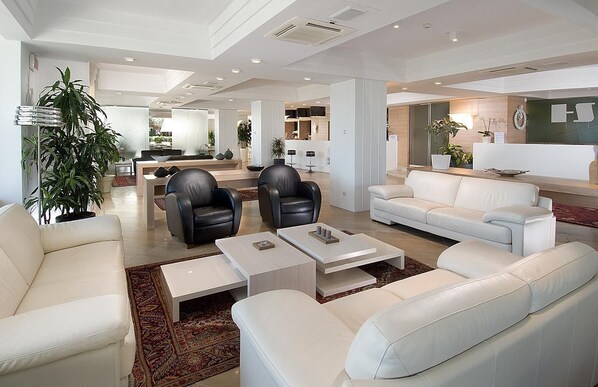 Una sala de estar moderna con sofás blancos, sillones de cuero negro y una mesa de centro. Se observa una planta grande en maceta y un área de cocina al fondo.