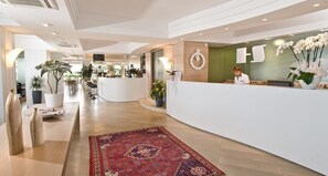Una moderna zona de recepción de hotel con un mostrador curvo, un recepcionista y diversas plantas en macetas.