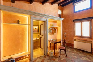 A bathroom with a sink, mirror, and toilet. There is a table and chair in the room.
