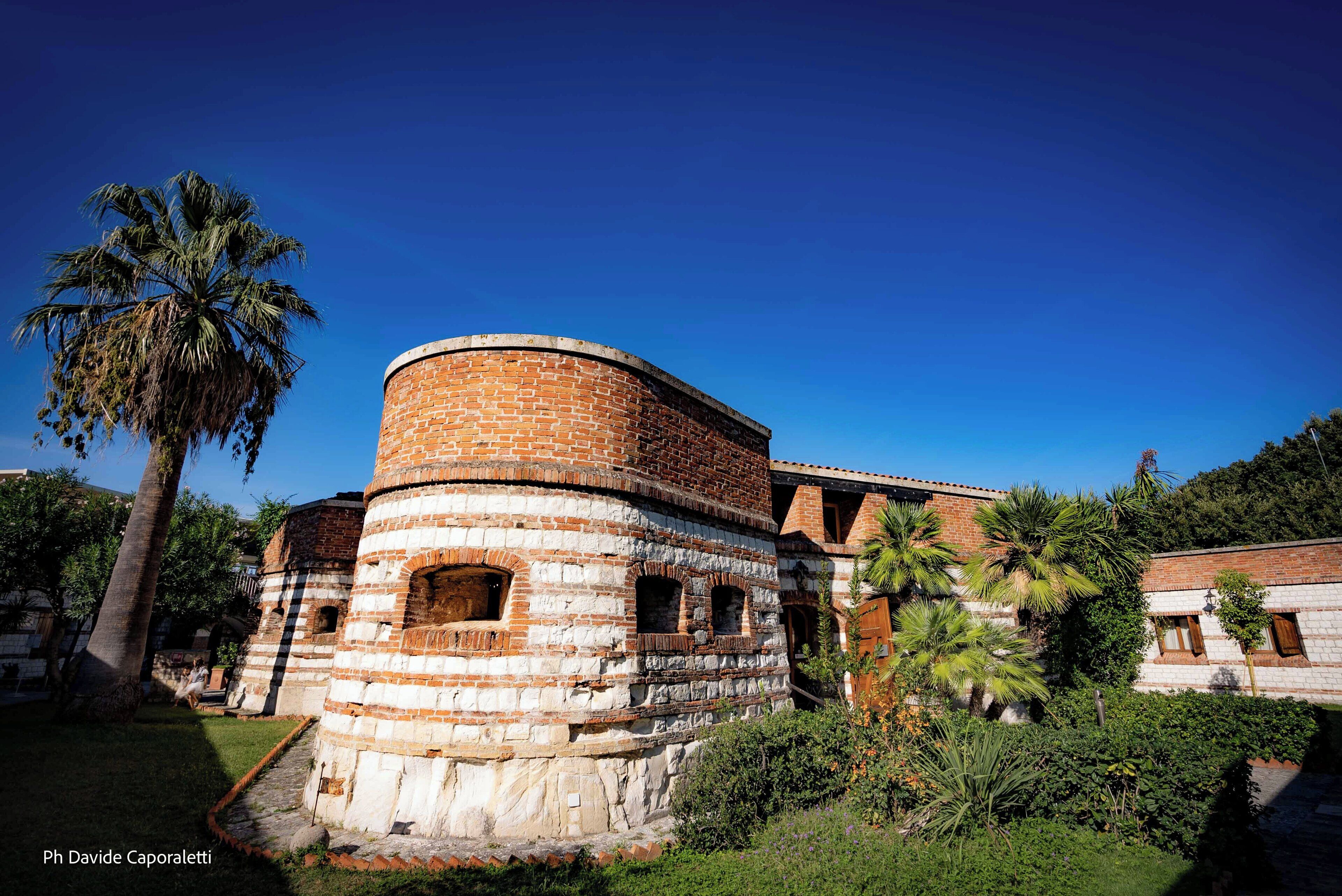 A historic brick building with a round tower, surrounded by lush greenery and palm trees.