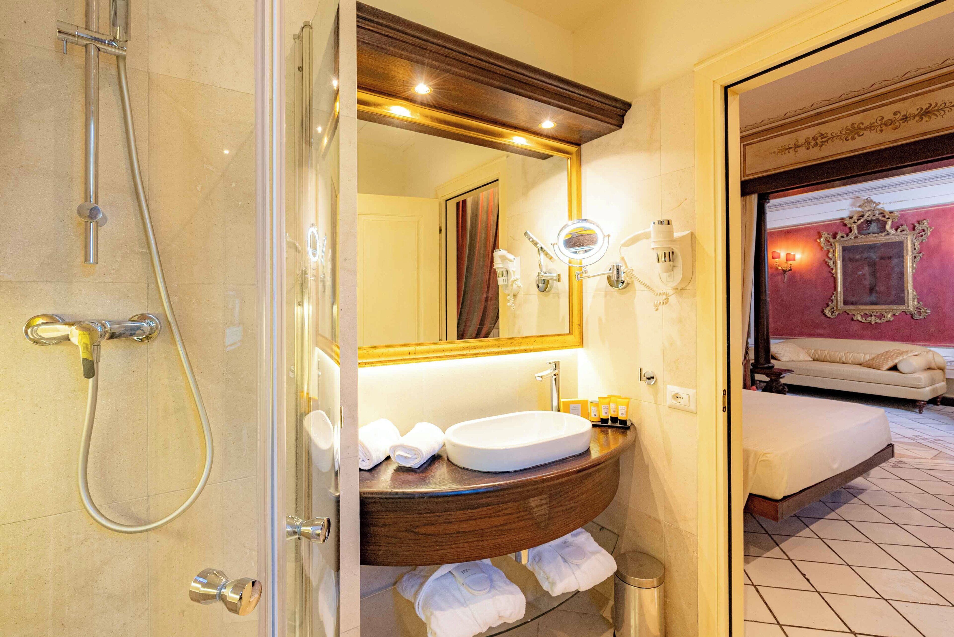 Ein Badezimmer mit Dusche, Waschbecken und Spiegel. Ein Bett ist im angrenzenden Raum zu sehen.