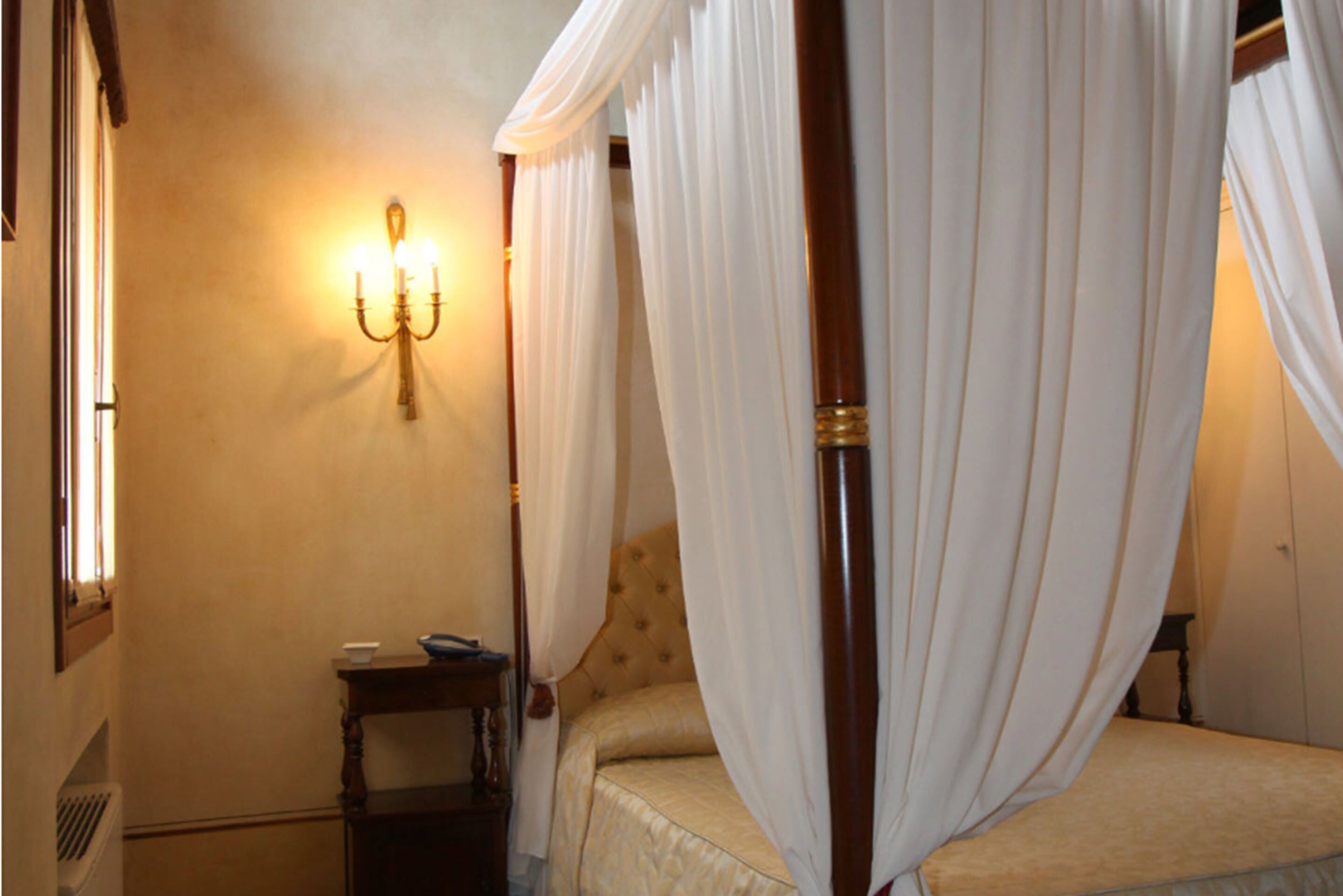 A bedroom with a four-poster bed, a bedside table, and a wall-mounted light fixture.