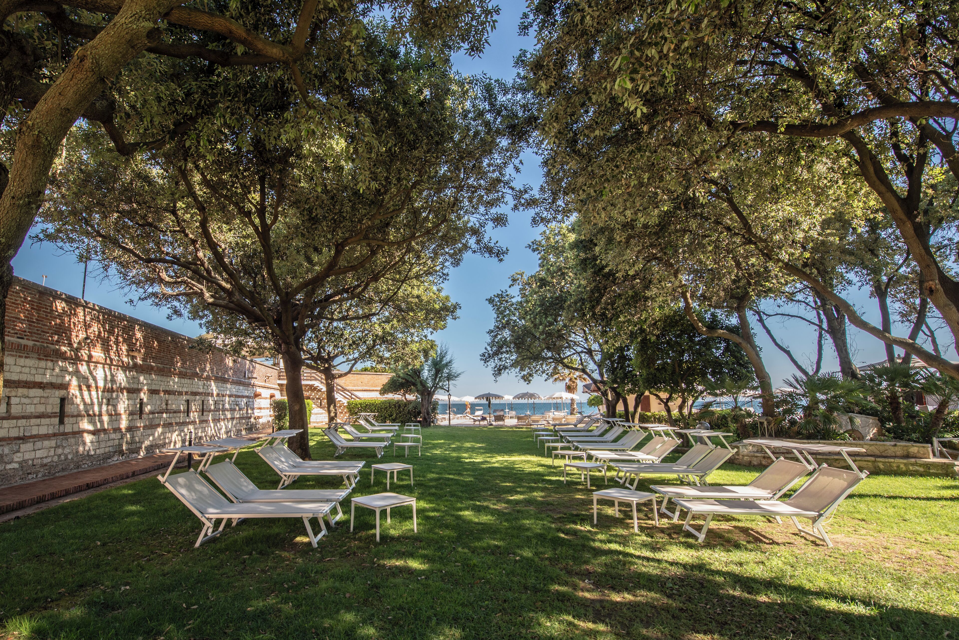 An outdoor seating area with sun loungers arranged in rows, a brick wall, and a view of the sea.