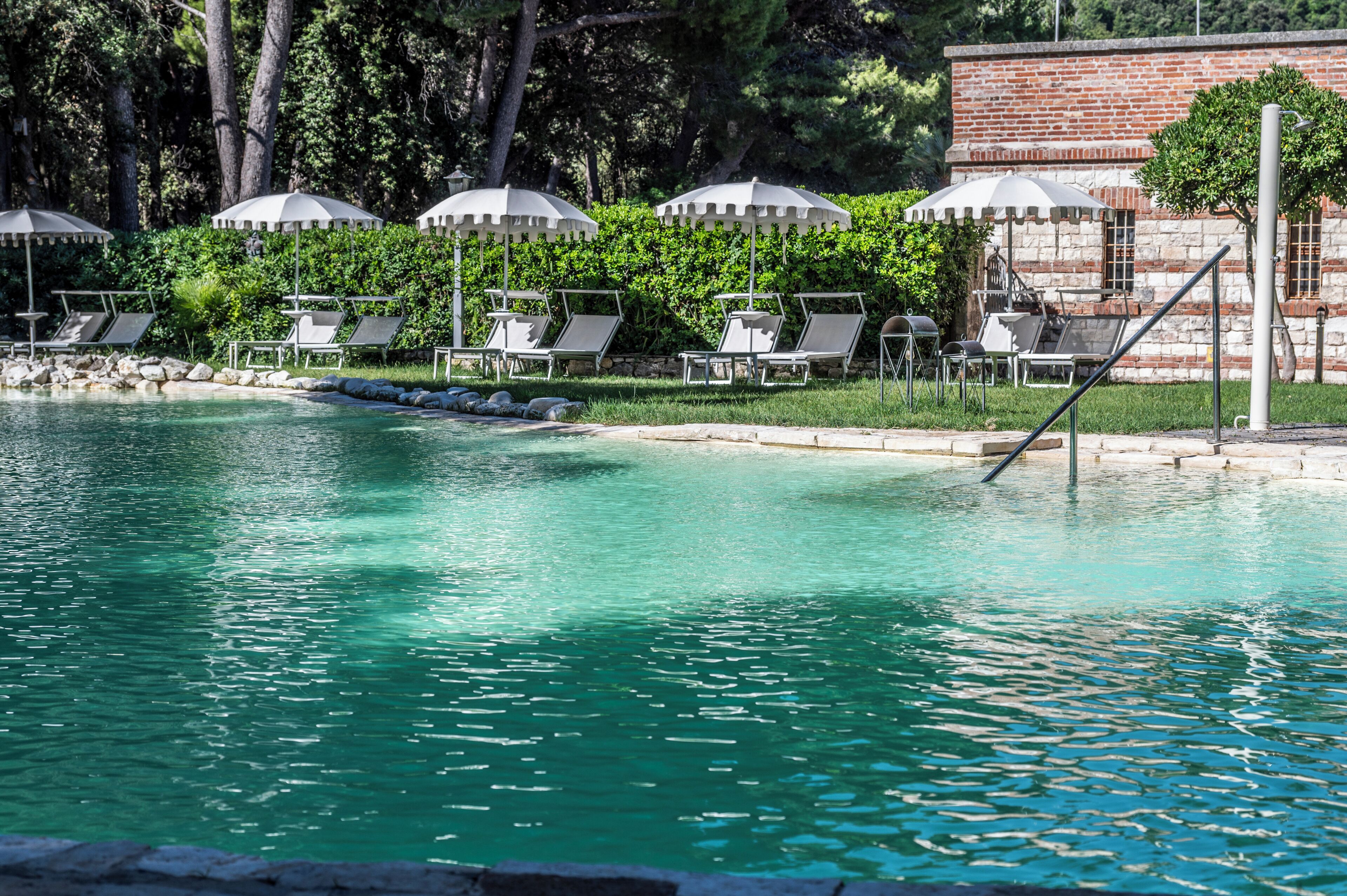 A pool with clear water, surrounded by lounge chairs and umbrellas, with a brick building in the background.