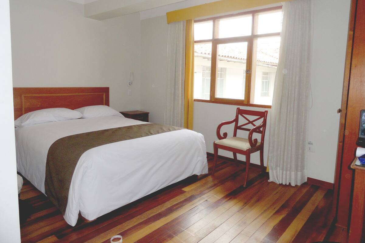 Habitación de hotel con piso de madera, cama con ropa de cama blanca, mesita de noche de madera, una silla y una ventana con cortinas.