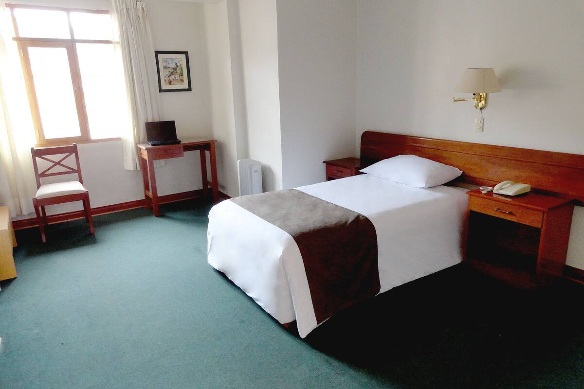 Una habitación de hotel con cama, mesita de noche, lámpara, silla y ventana con cortinas.