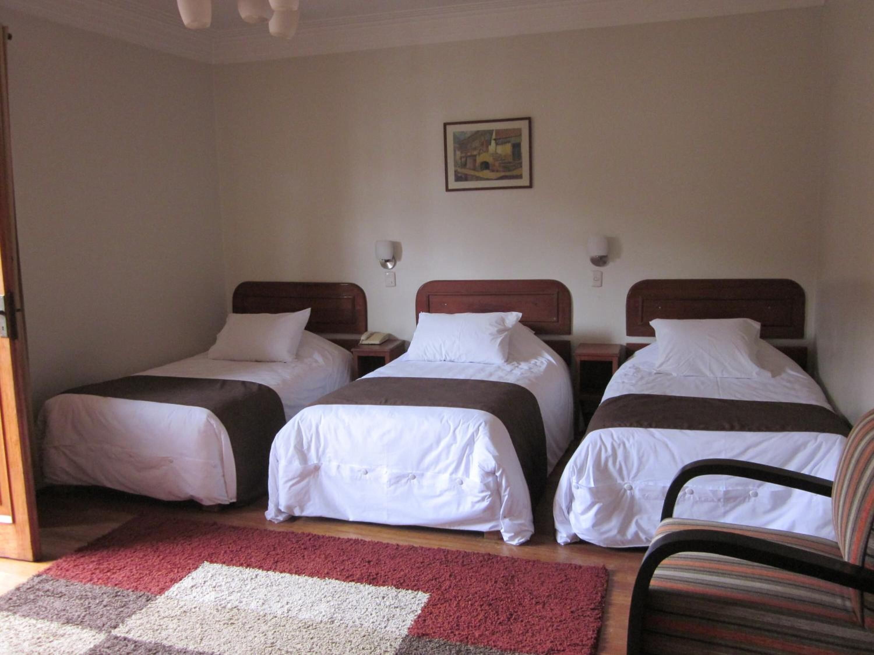 A room with three single beds, each with a wooden headboard and white bedding. There is a red and white rug on the floor, a chair with a striped cushion, and a painting on the wall.