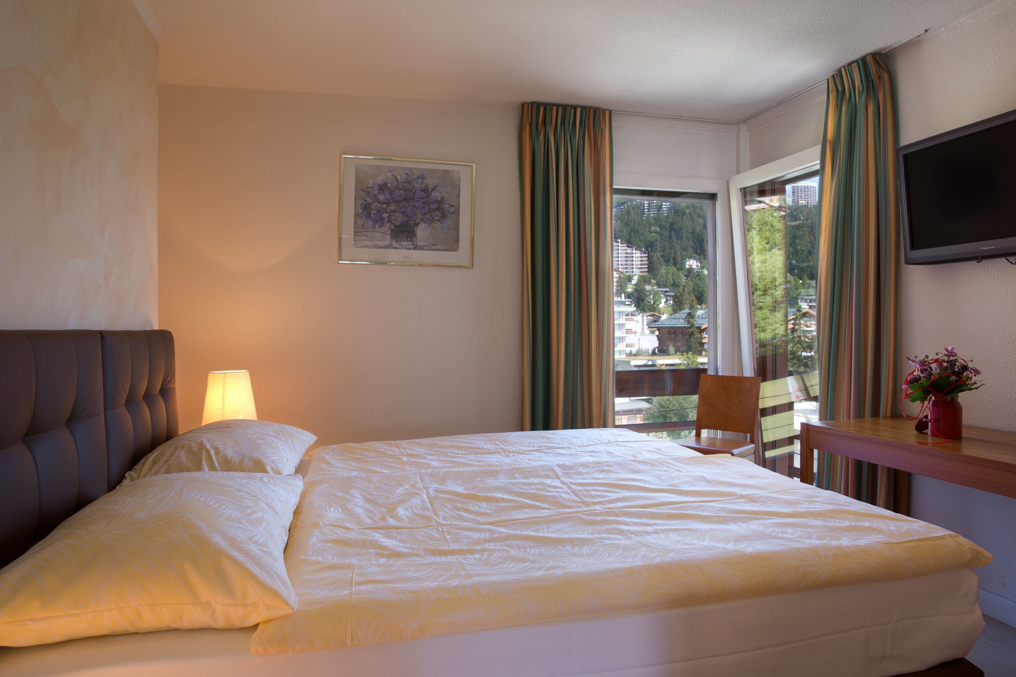 A hotel room with a bed, a chair, a table, a TV, and a window with curtains.