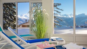 Habitación soleada con vistas a montañas nevadas, una planta y una mesa con un libro y una botella.