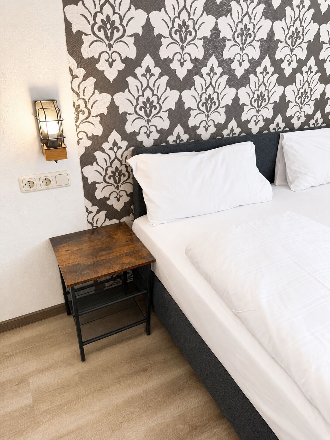 Une chambre d’hôtel bien aménagée, avec un lit, une table de chevet, une lampe fixée au mur et des prises électriques.