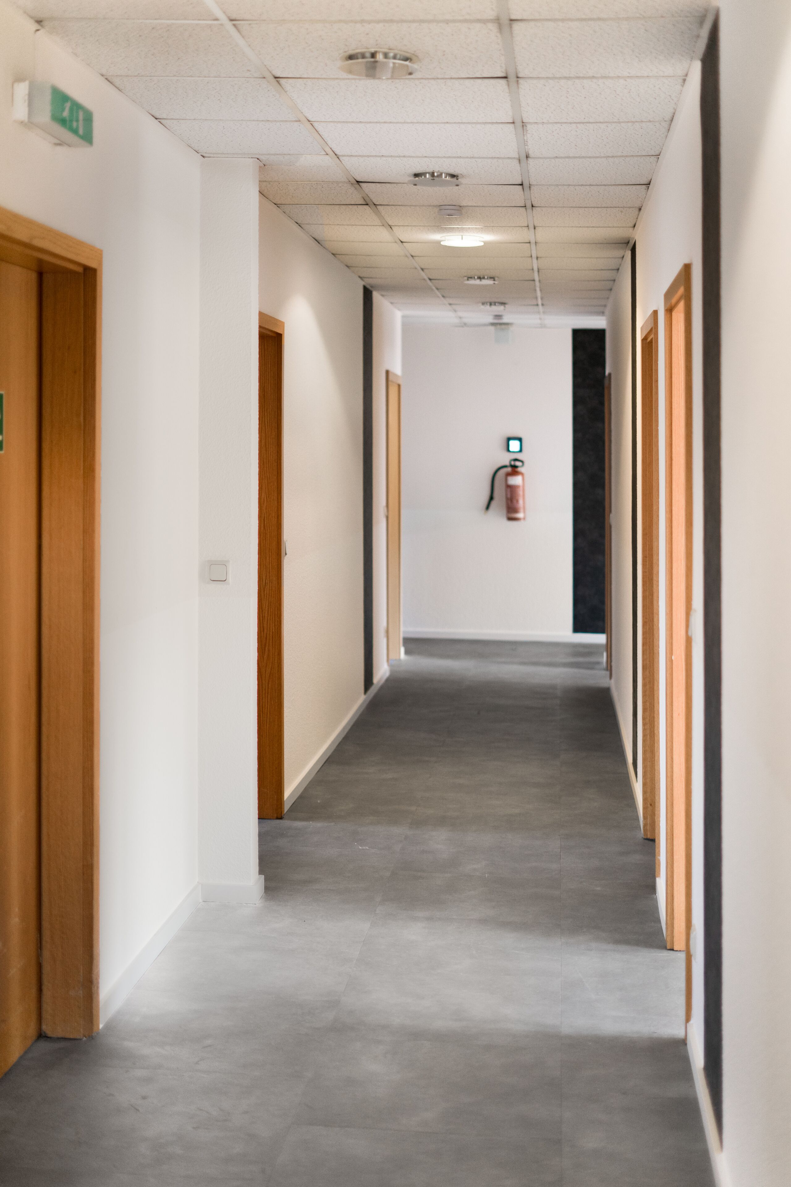 Een gang met houten deuren, een brandblusser aan de muur en een plafond met inbouwverlichting.