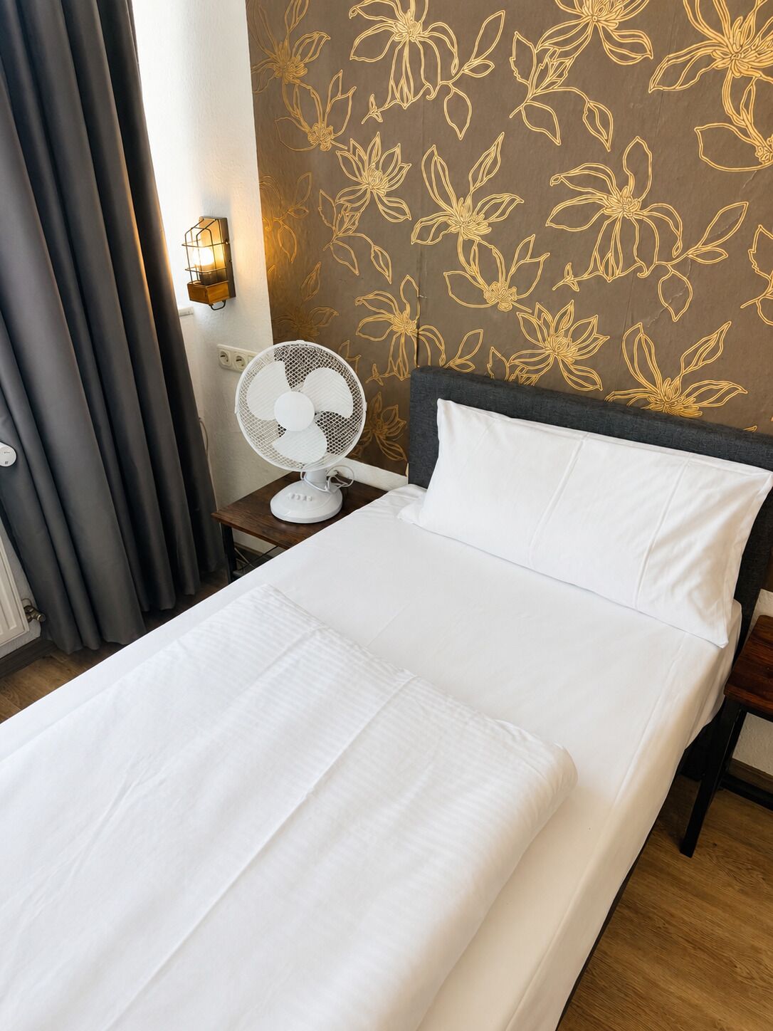 Habitación de hotel con cama, ventilador blanco, mesita de noche, lámpara y pared con estampado floral.