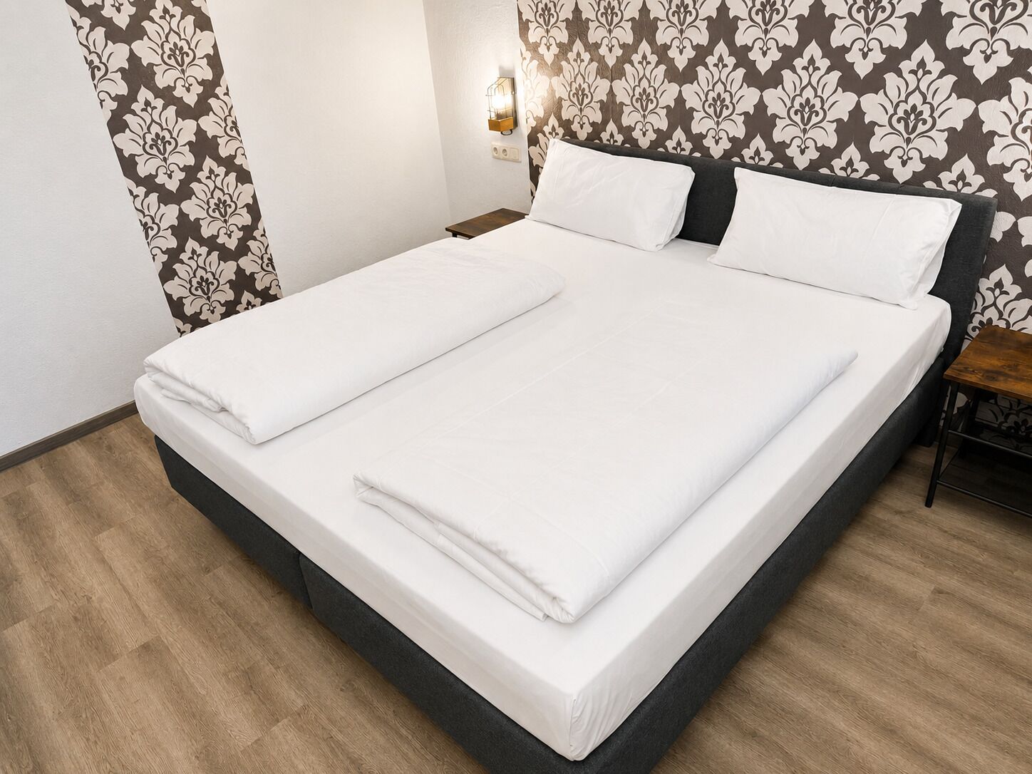 Ein ordentlich bezogenes Bett mit weißen Leinentüchern, einem dunklen Kopfteil und Tapete mit Muster an der Wand.