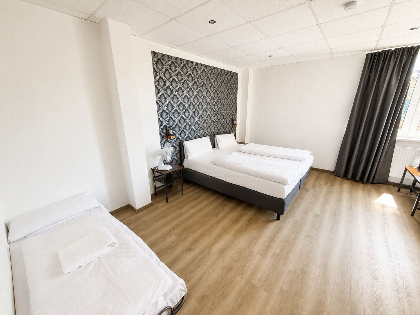 A hotel room with a large bed, a smaller bed, a bedside table, a wall-mounted heater, and a window with curtains.