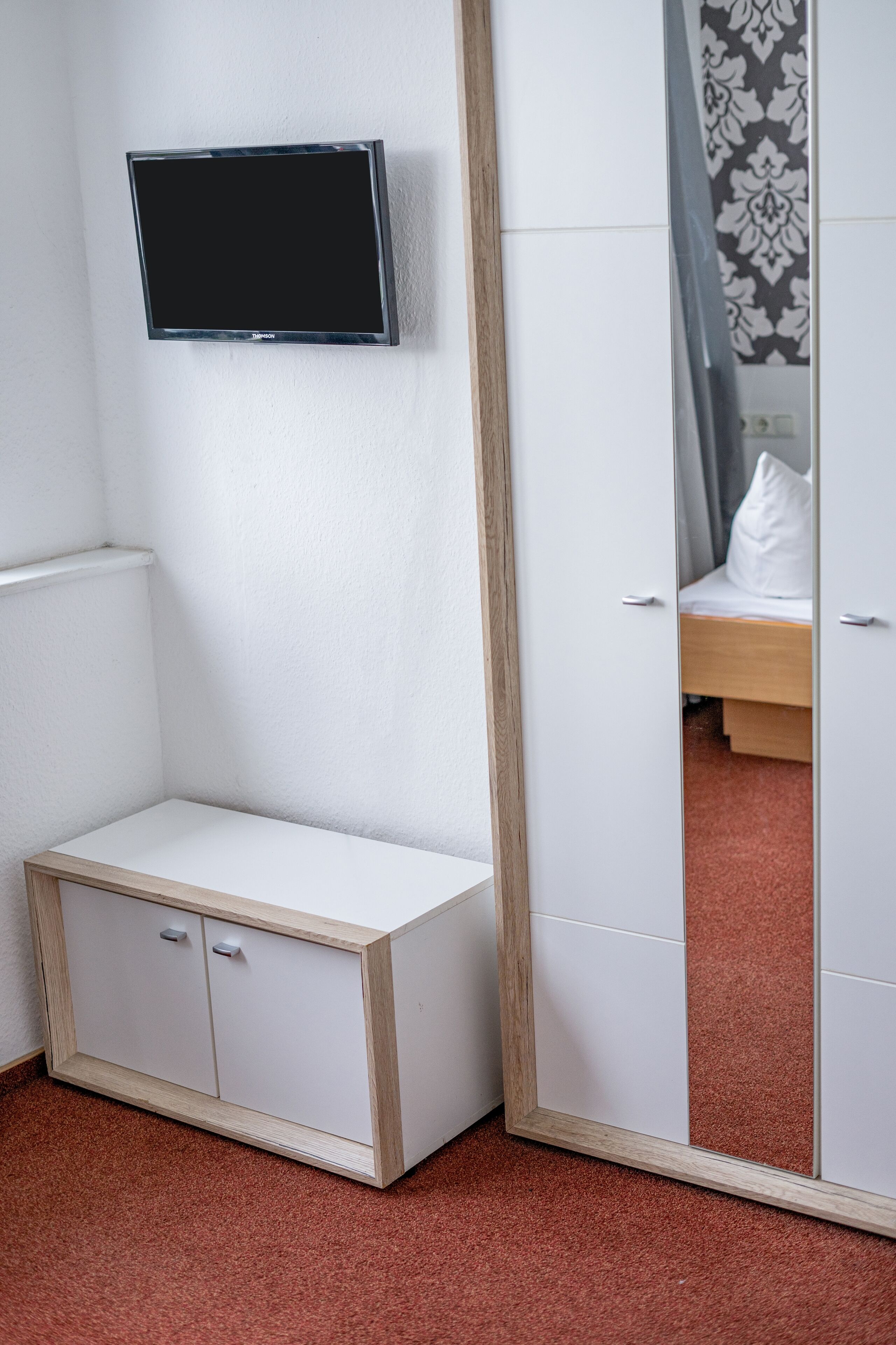 Una habitación con un armario blanco, un mueble blanco pequeño, un televisor montado en la pared y un espejo que refleja una cama y cortinas.