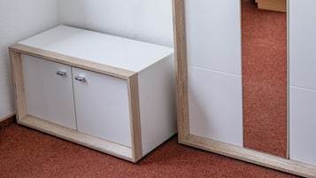 Quarto com um guarda-roupa branco, um pequeno armário branco, uma televisão na parede e um espelho refletindo uma cama e cortinas.