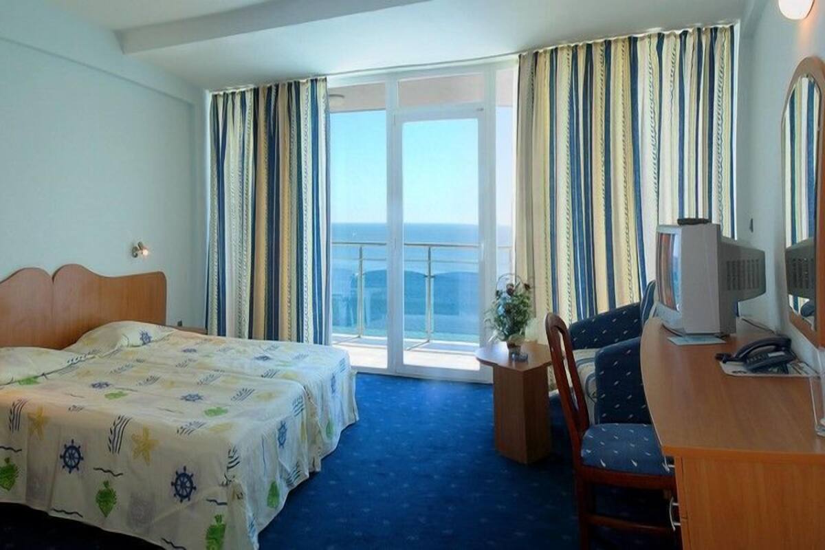 A hotel room with a bed, a desk, a chair, a television, and a view of the sea through large windows.