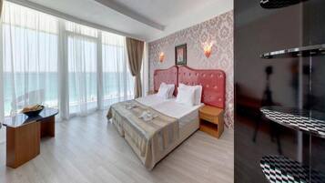 Ein Hotelzimmer mit einem großen Bett, einem Holfnachtkästchen, einem Schreibtisch mit Stuhl und einem Balkon mit Vorhängen.