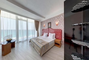 A hotel room with a large bed, a wooden nightstand, a desk with a chair, and a balcony with curtains. - Sirius Beach (Varna)
