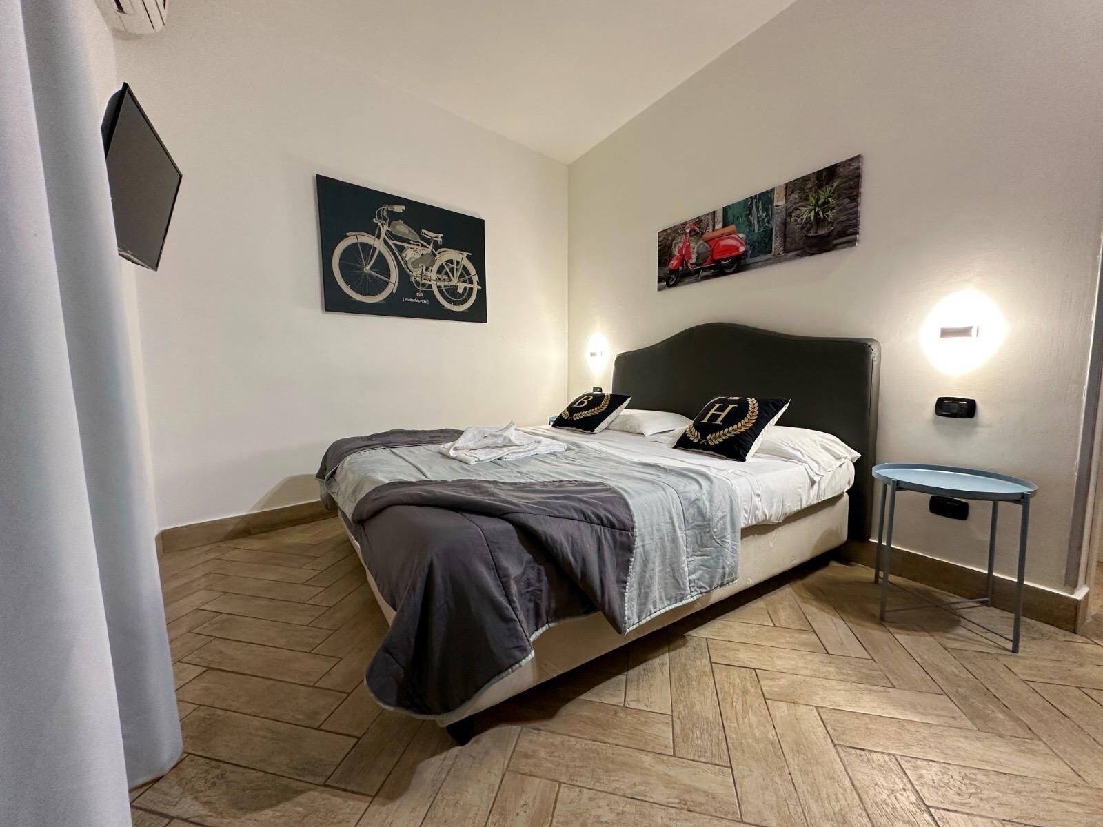 A neatly arranged bedroom with a bed, two wall-mounted lights, a small table, and two artworks on the wall.