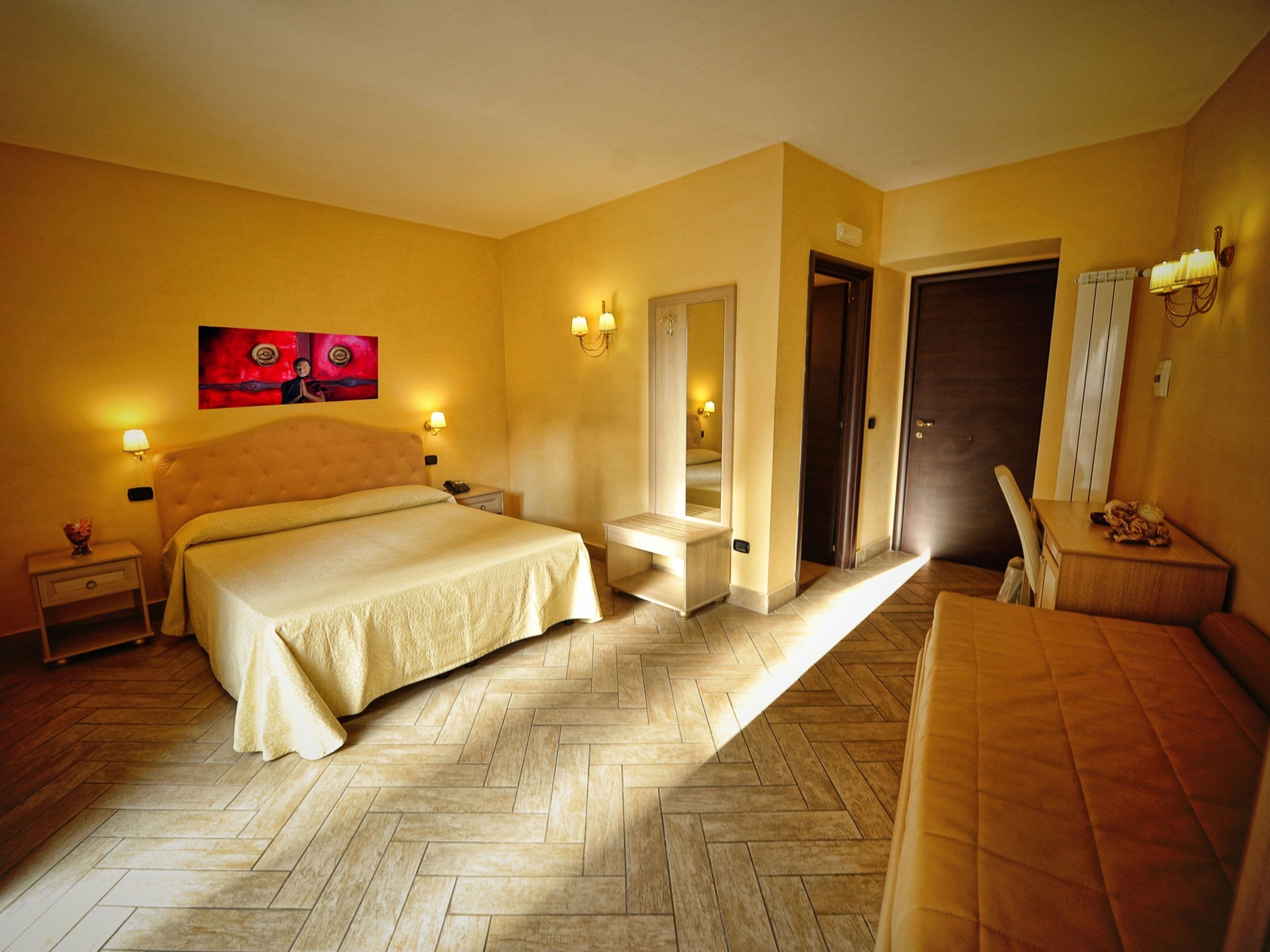 Habitación de hotel con una cama grande, mesitas de noche, un espejo, una puerta y una pequeña mesa con lámpara.
