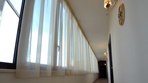 A hallway with a wooden floor, white walls, and a large window with sheer curtains. There is a wall-mounted light fixture and a door at the end of the hallway. - Hotel Barbato (Naples)