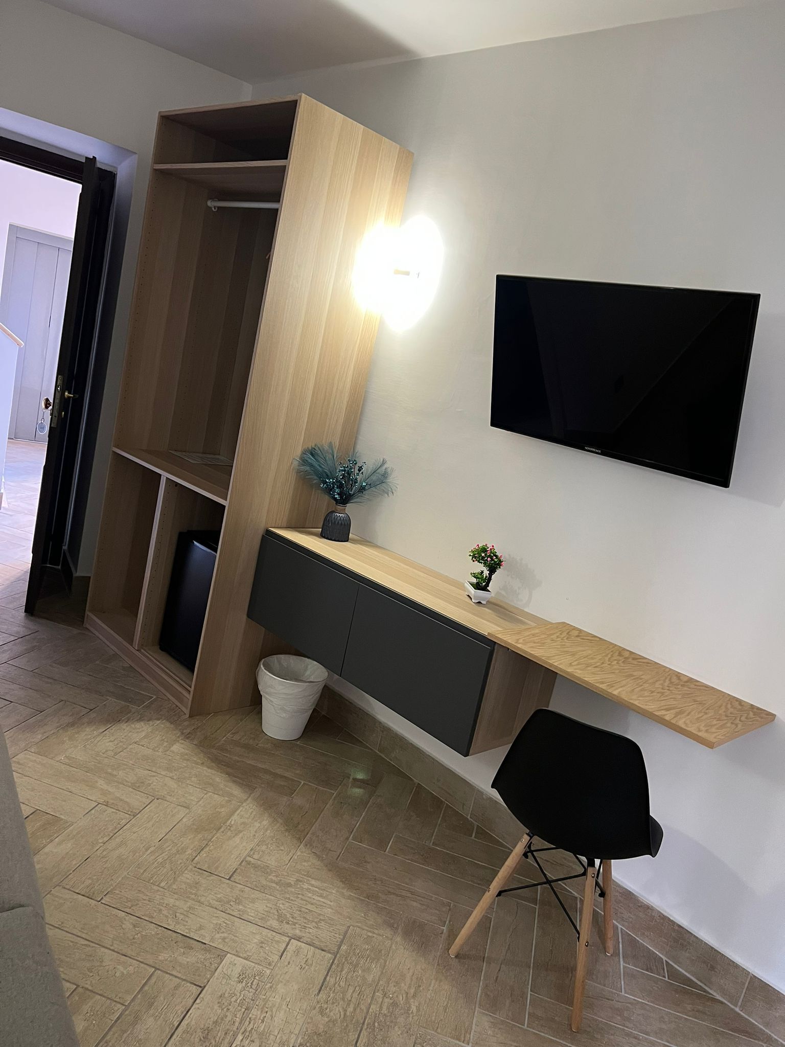 A modern room with a wooden wardrobe, a wall-mounted TV, a small desk with a plant, and a black chair.