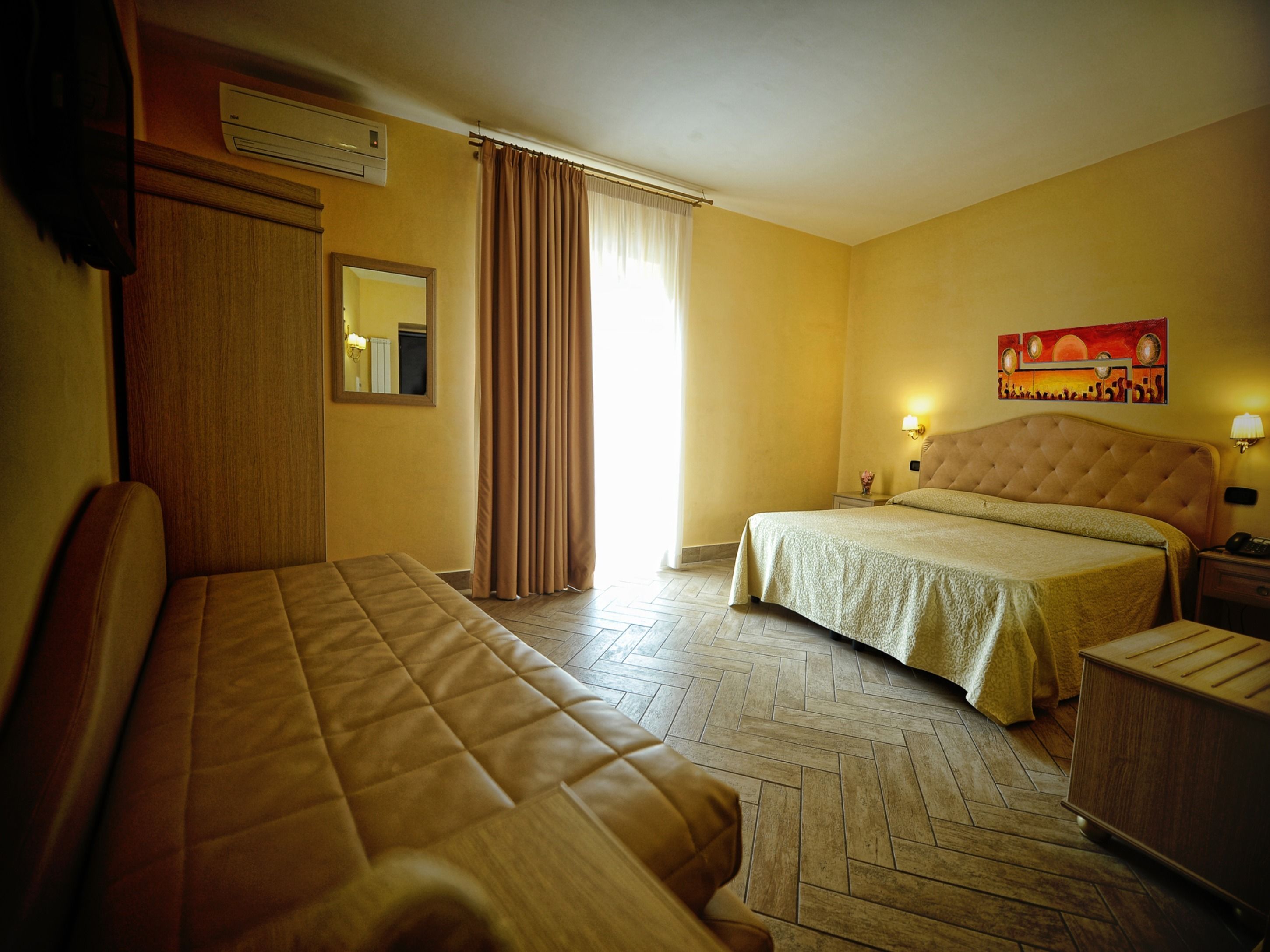 A hotel room with two beds, a wooden floor, a mirror, a painting, and a nightstand with a lamp.