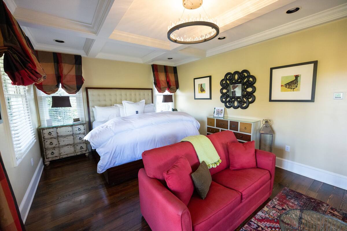 A bedroom with a large bed, a red sofa, a chandelier, and artwork on the walls.