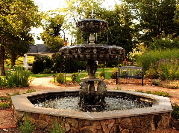 A multi-tiered fountain with water cascading down, surrounded by a stone basin and set in a garden with various plants and a bench. - Inn at Willow Grove (Orange)