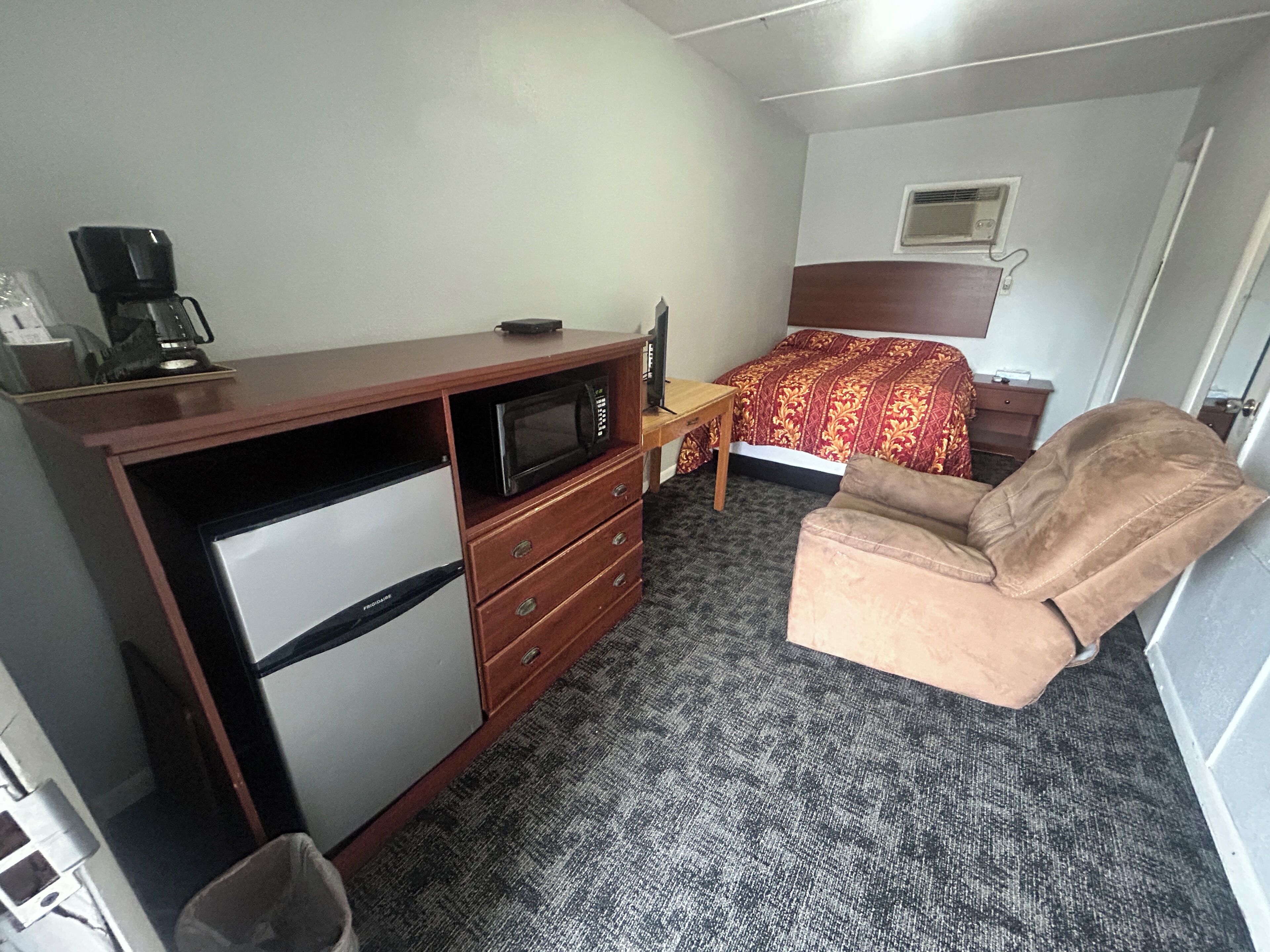Een compacte hotelkamer met kitchenette, een bed met een gestreept beddegoed, een bruine fauteuil en een klein bureau met een magnetron.