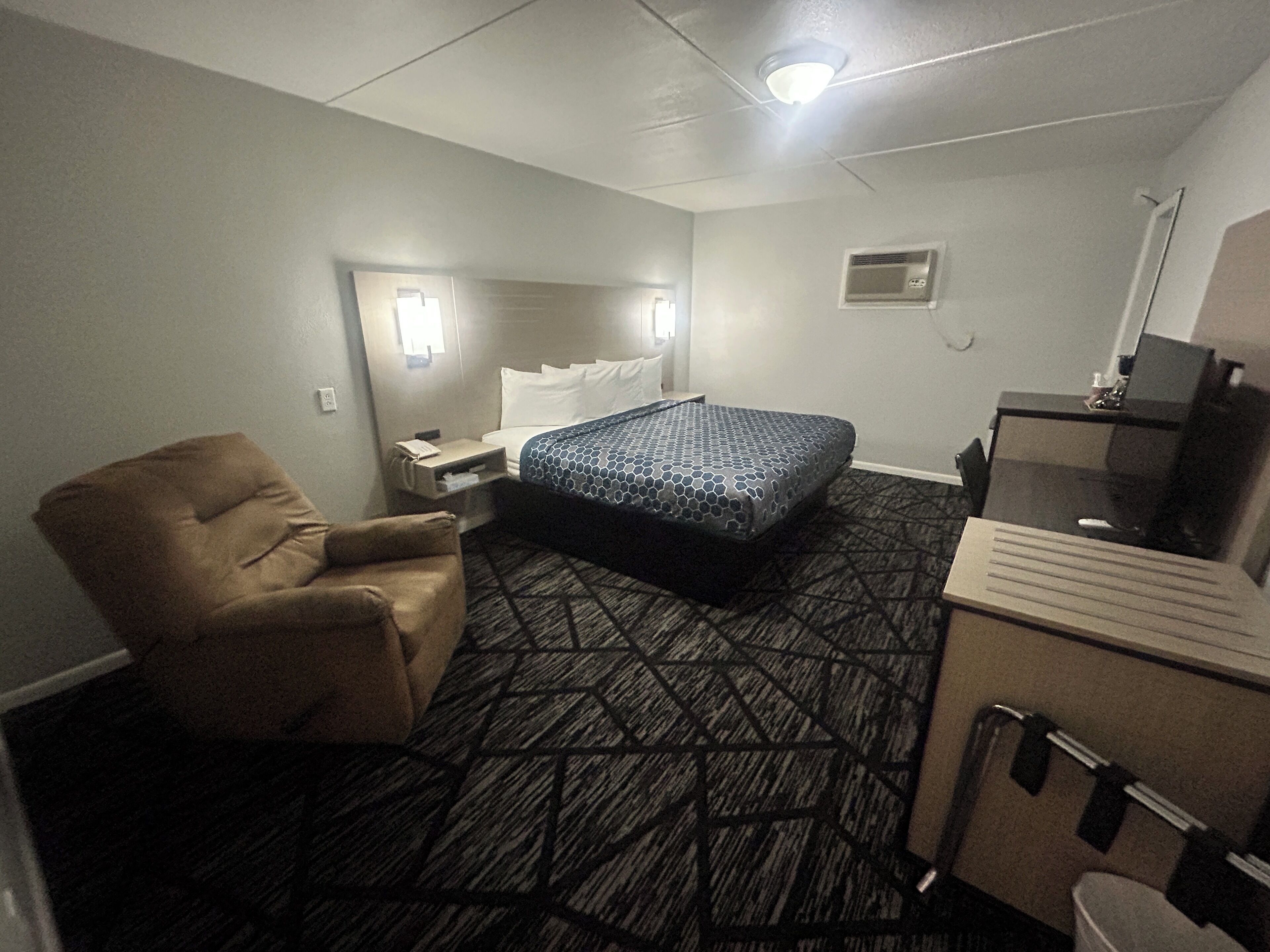 A hotel room with a bed, a chair, a desk, a mirror, and a small kitchenette.