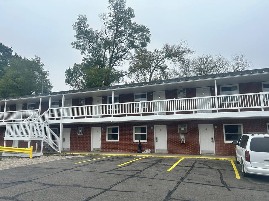 Um motel de dois andares com uma escadaria central, um estacionamento com linhas amarelas e um carro branco estacionado no local.