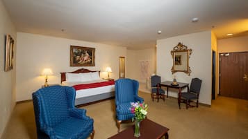 Una habitación de hotel con una cama, dos sillones azules, una mesa de centro de madera con un jarrón de flores, una mesita con una lámpara y un espejo en la pared.