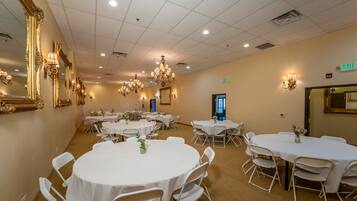 A spacious banquet hall with multiple round tables and chairs, elegant chandeliers, and wall-mounted lighting fixtures.