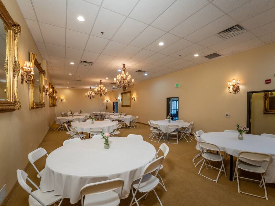 A spacious banquet hall with multiple round tables and chairs, elegant chandeliers, and wall-mounted lighting fixtures.