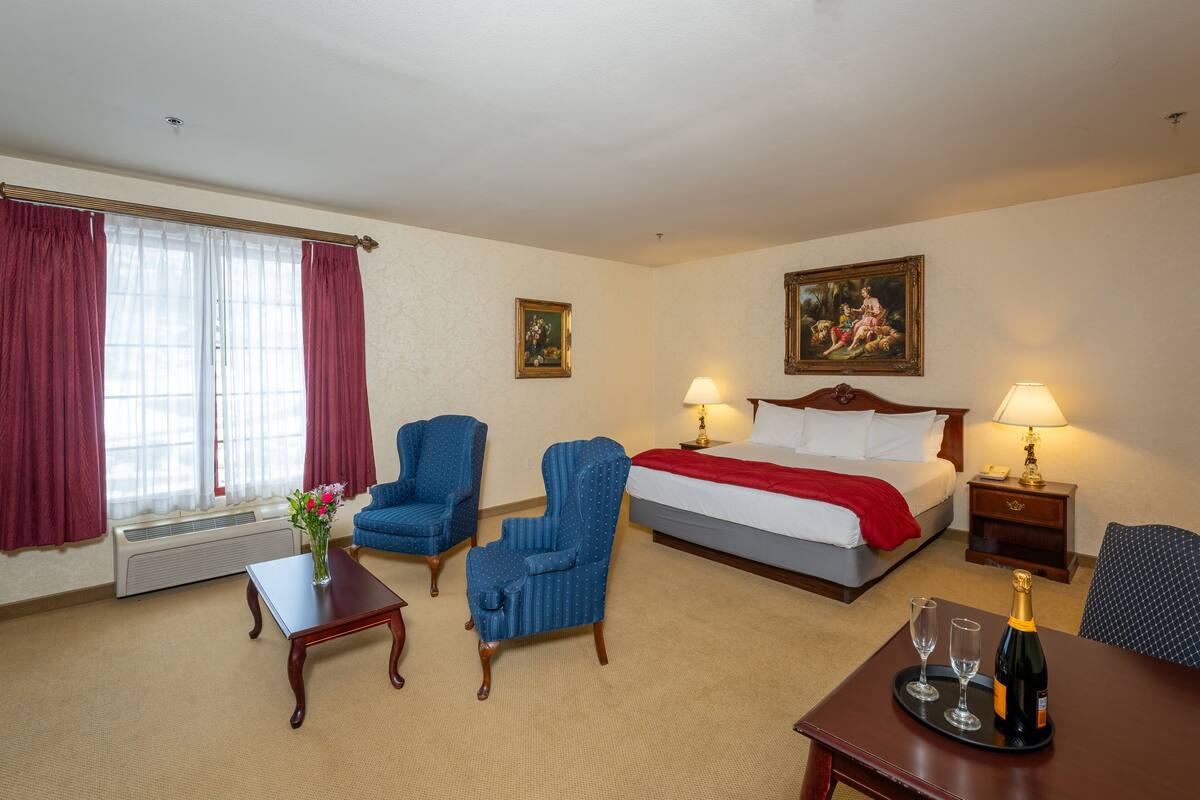 A hotel room with a bed, two chairs, a small table with a vase of flowers, a bedside table with a lamp, and a painting on the wall.
