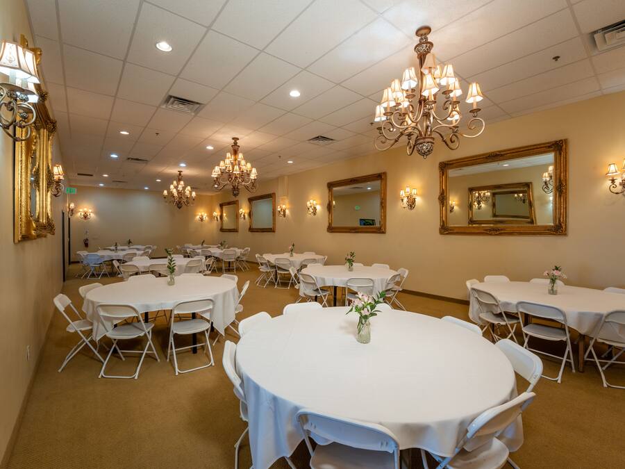 Uma sala de banquetes com mesas redondas, cadeiras brancas, lustres, arandelas e espelhos.