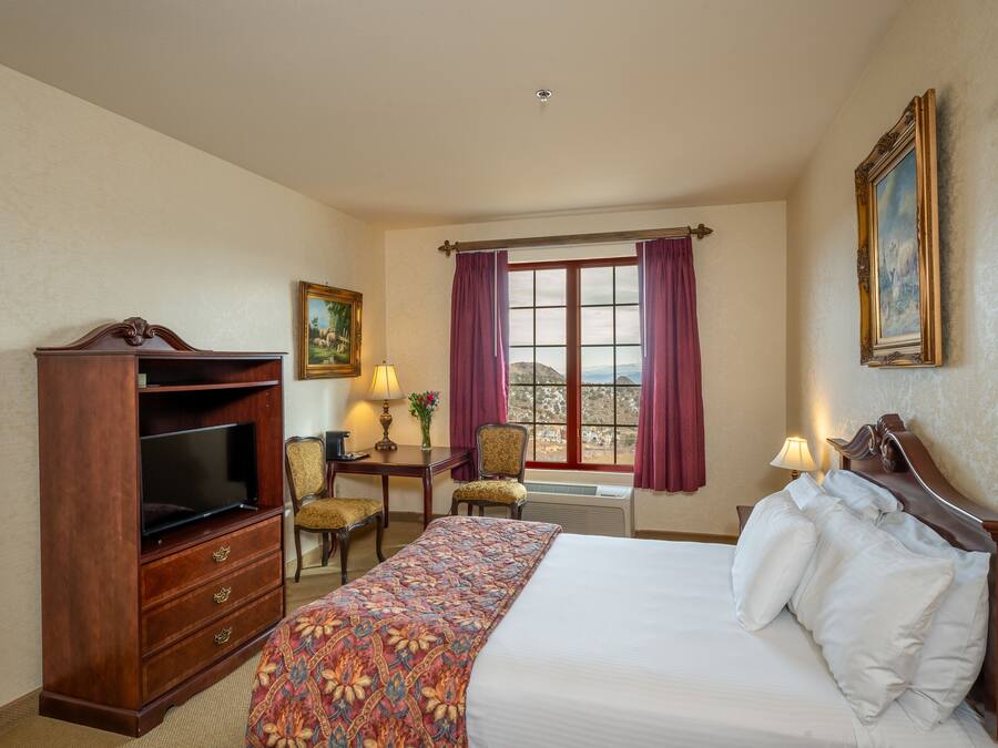 Quarto de hotel com cama, televisão, secretária com luminária e janela com cortinas.