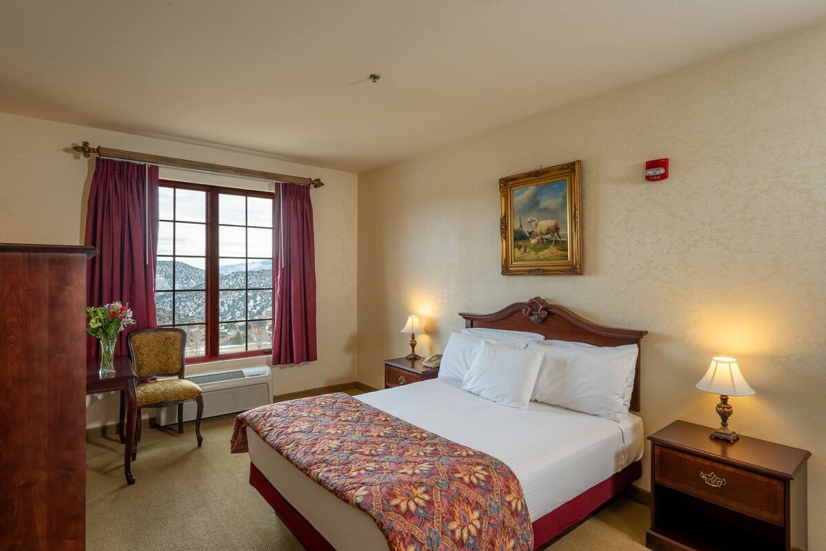 A hotel room with a bed, a chair, a desk, a window with curtains, a painting on the wall, and a nightstand with a lamp.