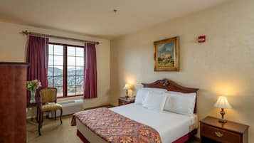 A hotel room with a bed, a chair, a desk, a window with curtains, a painting on the wall, and a nightstand with a lamp.