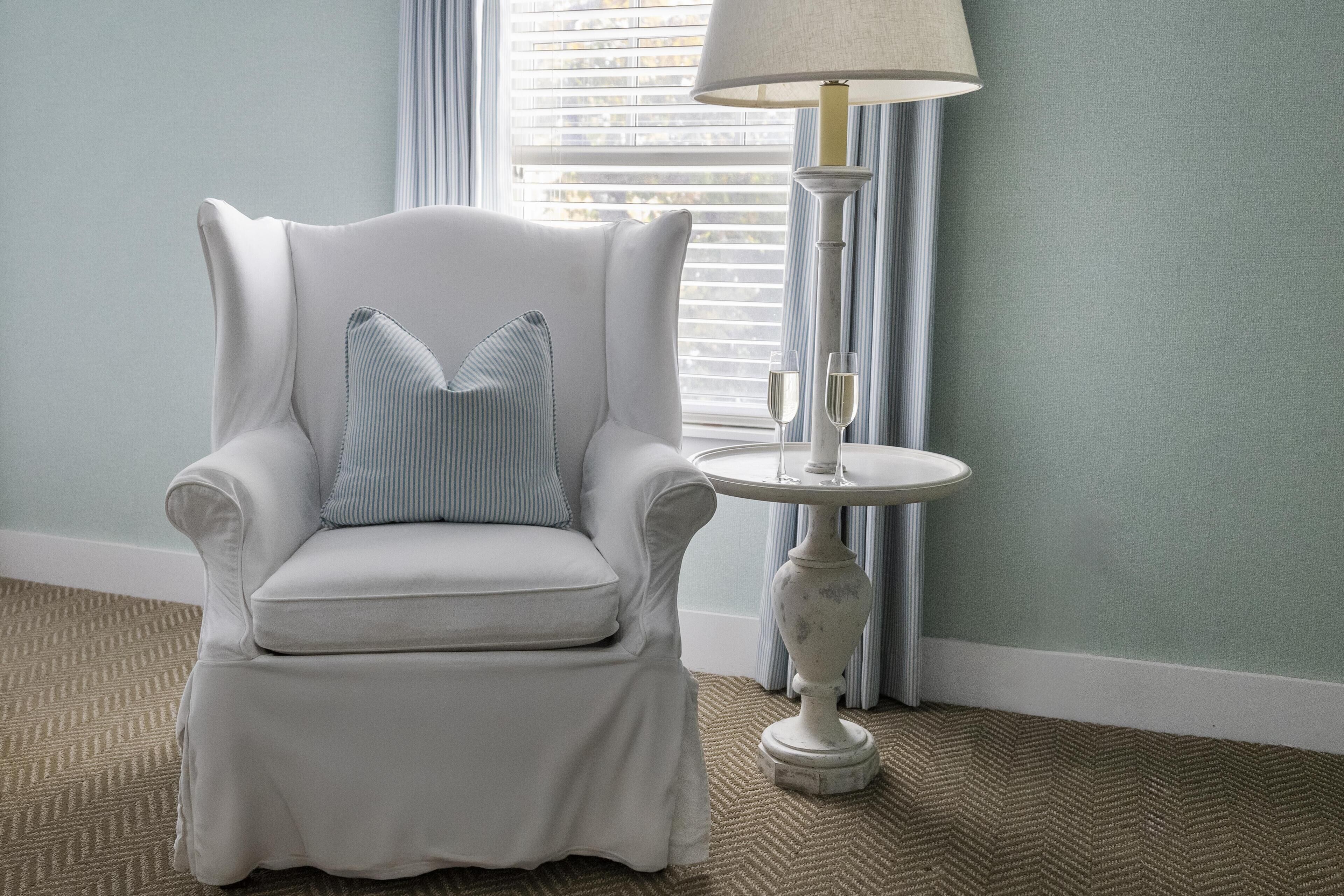 A white armchair with a striped pillow, a table with a lamp, and a window with blinds.