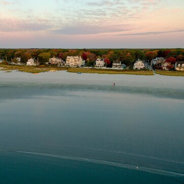 An aerial view of a waterfront property with a row of houses along the shore, a calm body of water, and a distant shoreline.