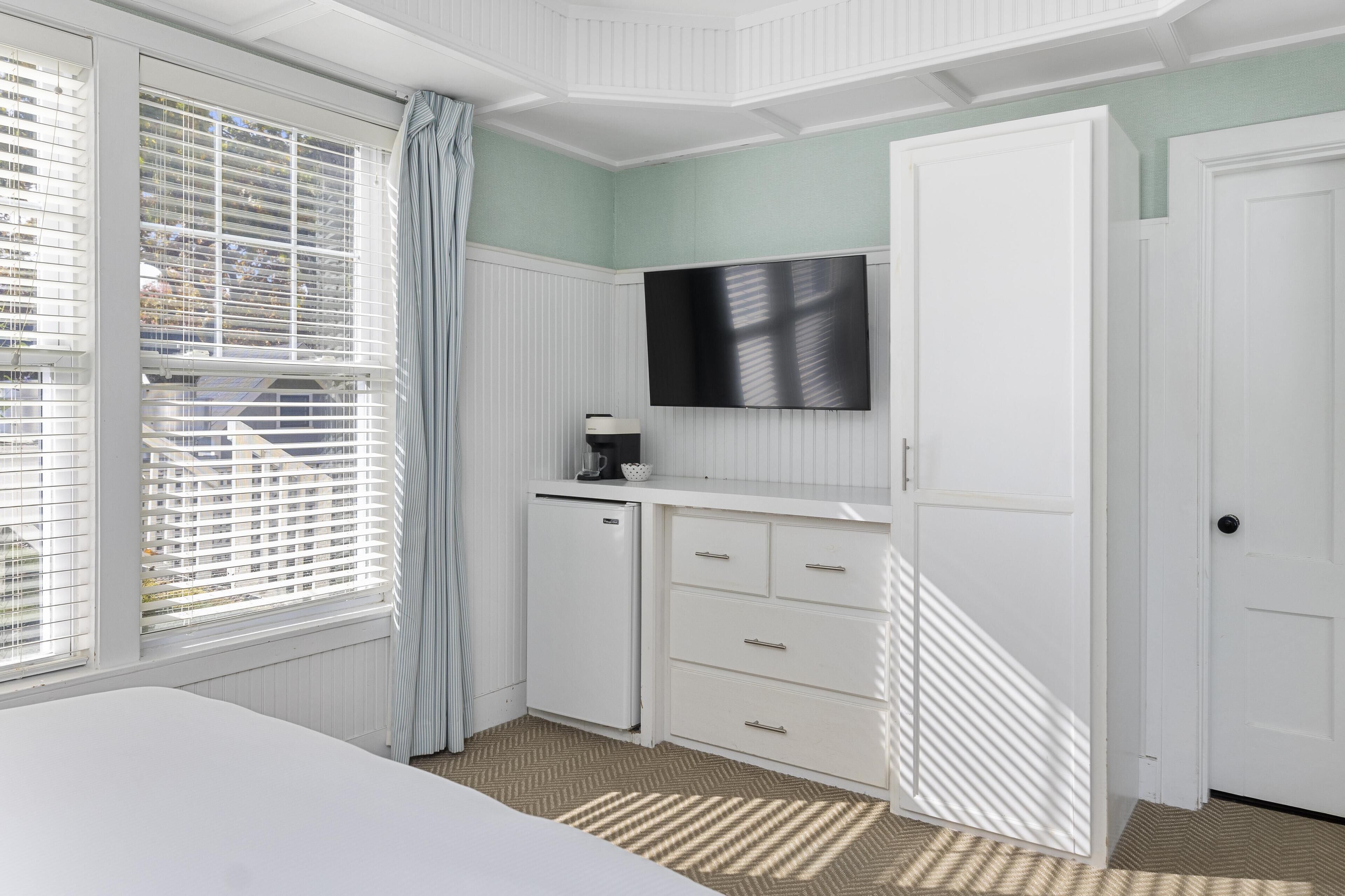A bedroom with a bed, a TV mounted on the wall, a small refrigerator, and a window with blinds.