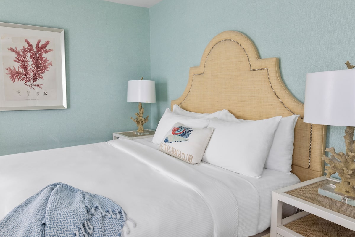 A neatly made bed with white linens, a headboard with a chain detail, and a framed botanical print on the wall.