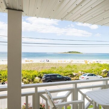 A covered balcony with a view of the beach, white outdoor furniture, and parked cars.